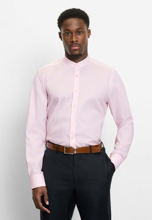 Mann trägt ein hellrosa Hemd mit Stehkragen, das in schwarze Hosen mit einem braunen Gürtel gesteckt ist, und steht vor einem schlichten weißen Hintergrund.