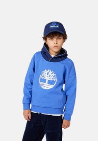 Jongen in een blauwe Timberland hoodie met een boomlogo, marineblauwe corduroy broek en een marineblauwe Timberland pet, staande tegen een effen achtergrond.