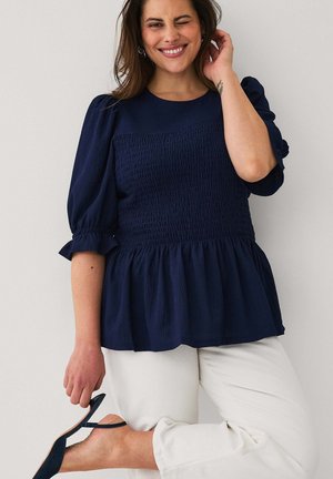 Kvinde i navy puff-ærmet top og hvide bukser blinker med et øje, mens hun holder en hæl tæt ved sin hævede fod mod en ensartet lys baggrund.