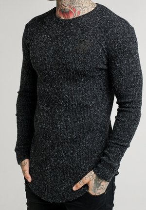 Mand iført en tætsiddende mørkegrå tekstureret strikket sweater med rund hals og subtilt broderet logo på brystet, hænderne i lommerne.