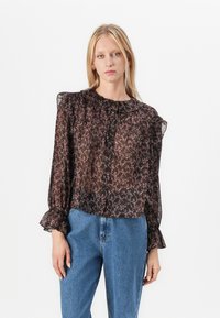 Blusa nera trasparente con motivo floreale, con scollatura volant e chiusura a bottoni. Presenta maniche lunghe con polsini svasati. Indossata sopra jeans in denim blu.