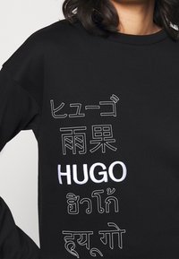 Sudadera negra con la palabra "HUGO" escrita en letras blancas y contorneadas en múltiples escrituras asiáticas en la parte frontal, llevada por una persona de cabello oscuro.