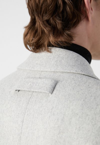 Manteau en feutre gris avec un col cranté, une texture subtile et une languette cousue avec le logo "HUGO" au dos.