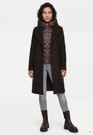 Standing woman wearing a dark long coat over a brown quilted jacket, light gray skinny jeans, and dark chunky boots against a white background.