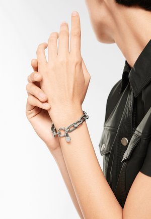 Zilveren kettingarmband met een klein hangslotbedel, voorzien van in elkaar grijpende schakels en een gladde textuur, gedragen op een hand tegen een effen achtergrond.
