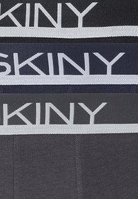 Fasce di tessuto grigie, blu navy e grigie scure impilate con il testo bianco "SKINY" sui cinturini elasticizzati della biancheria intima.