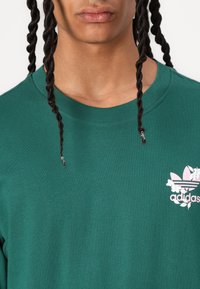Zaļš kokvilnas T-krekls ar apaļu kaklu, īsām piedurknēm un rozā ziedu Adidas logo kreisajā krūšu daļā.