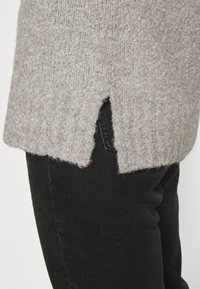 Pull en tricot gris clair avec une finition texturée et des fentes sur les côtés, associé à un jean noir. Le pull a une coupe décontractée et ample.