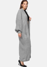 Lungo cardigan grigio lavorato a maglia, oversize con maniche ampie, dotato di una tasca frontale; indossato su una maglietta nera e abbinato a tacchi neri.