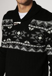 Pull en tricot noir avec un motif géométrique en gris et blanc, doté d'un col châle et d'une fermeture à toggles au niveau du cou.