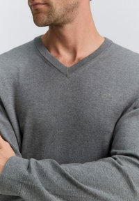 Pulover gri cu decolteu în V, confecționat dintr-un material moale. Prezintă un guler și manșete cu căptușeală, având un logo mic pe piept. Textură netedă.