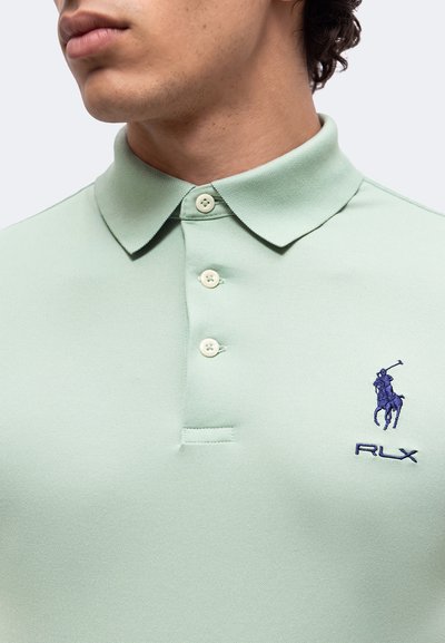 Homme portant un polo vert pâle avec trois boutons et un logo joueur de polo brodé bleu marine sur la poitrine.