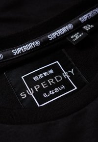 T-shirt en coton noir avec une étiquette au cou portant le logo "SUPERDRY" en blanc sur fond noir et une étiquette d'instructions de soin attachée.