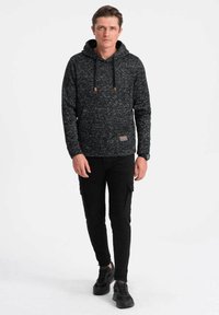 Zwarte, gestructureerde hoodie met een trekkoordkap en een patchdetail, gecombineerd met zwarte cargobroeken en zwarte sneakers.