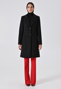 Una donna in piedi che indossa un cappotto nero con bottoni sopra un dolcevita nero, pantaloni rossi a gamba larga e scarpe nere a punta con punte dorate.