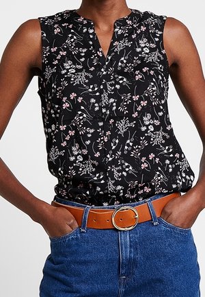 Mouwloze zwarte blouse met roze en witte bloemenprint, ingestopt in een blauwe spijkerbroek met een bruine leren riem met een ronde gouden gesp.