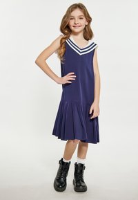Marineblaue ärmellose Kleid mit einem weißen Matrosenkragen und drei Streifen, das einen schwingenden, plissierten Rock hat. Kombiniert mit schwarzen Schnürstiefeln.