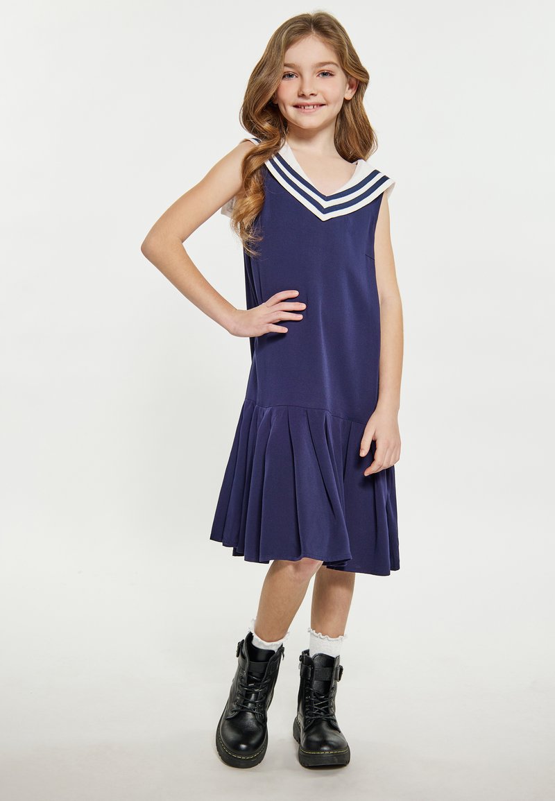 Marineblaue ärmellose Kleid mit einem weißen Matrosenkragen und drei Streifen, das einen schwingenden, plissierten Rock hat. Kombiniert mit schwarzen Schnürstiefeln.