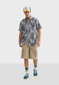 Košelia s krátkymi rukávmi a gombíkmi s modro-bežovým vzorom listov, skombinovaná s kaki cargo šortkami, farebnými ponožkami a teal topánkami.