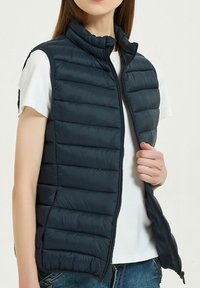 Gilet rembourré bleu marine avec un design à matelassage vertical, un col montant et une fermeture éclair intégrale. Porté par-dessus un t-shirt blanc, il présente une texture lisse et douce.