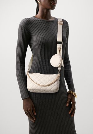 Donna che indossa un abito scuro a maniche lunghe a coste con una borsa a tracolla trapuntata color crema e un portamonete rotondo abbinato attaccato alla tracolla.