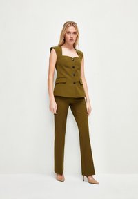 Olijfgroene mouwloze blouse met vierkante halslijn, vijf zwarte knopen en zakken, gecombineerd met bijpassende fitted broek en tan hoge hakken.