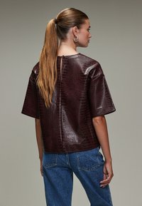 Burgundy faux leather top with a croc texture, short sleeves, and a back button closure. Paired with light blue denim pants.