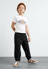White T-shirt with blue and orange "Dickies" logo, black sweatpants, and brown, white, and black sneakers. Clear background and neutral pose.