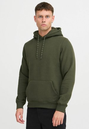 Green hoodie made of textured fabric, featuring a front pocket, adjustable drawstrings, and ribbed cuffs. Model is wearing black pants.