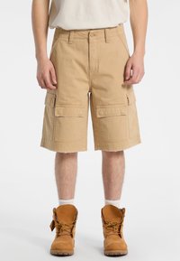 Shorts cargo beiges avec une coupe décontractée, présentant deux grandes poches latérales, un ourlet effiloché, et fabriqués en tissu en coton durable. Botte de travail marron visible.