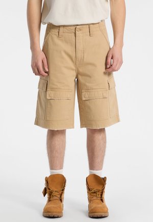 Beige Cargo-Shorts mit lockerer Passform, ausgestattet mit zwei großen Seitentaschen, fransigem Saum und aus strapazierfähigem Baumwollstoff gefertigt. Braune Arbeitsstiefel sichtbar.