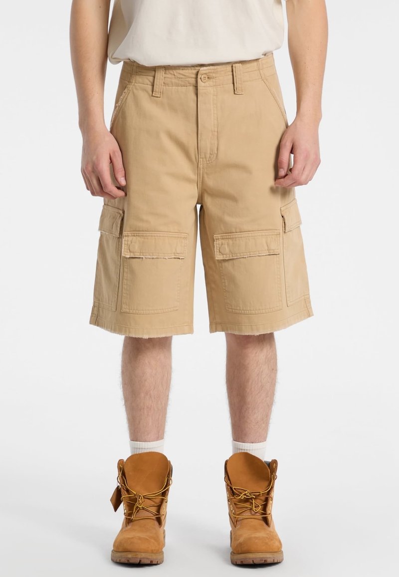Shorts cargo beiges avec une coupe décontractée, présentant deux grandes poches latérales, un ourlet effiloché, et fabriqués en tissu en coton durable. Botte de travail marron visible.