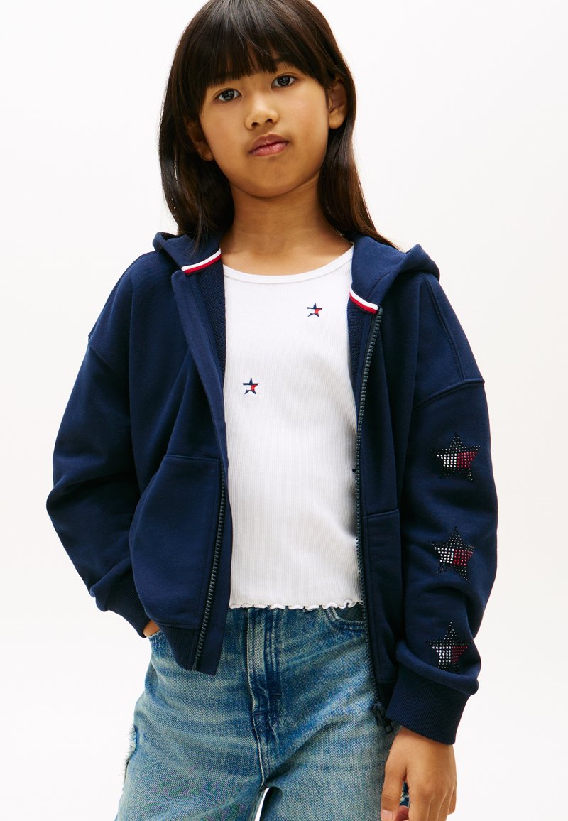 Νεαρή κοπέλα που φοράει μπλε σκούρα ζακέτα με φερμουάρ και σχέδια αστεριών στο μανίκι, λευκό αμάνικο τοπ με μικρά σχέδια αστεριών και τζιν παντελόνι.