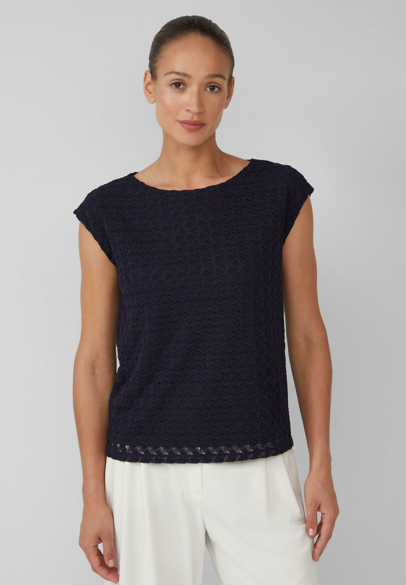 Haut bleu marine à manches courtes avec un motif géométrique texturé, un col rond et une coupe légèrement ample, associé à un pantalon de couleur claire.