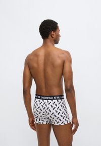 Vyriški boksininkų šortai su juodu elastiniu juosmeniu, ant kurio parašyta „KARL LAGERFELD“, balta baze ir besikartojančiu juodu grafiniu raštu.