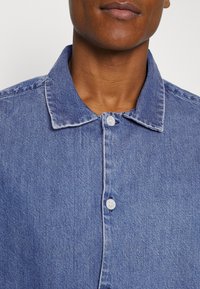 Gros plan sur une personne portant une chemise en jean bleue avec des boutons blancs et un col pointu.
