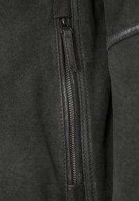 Schwarze Lederjacke mit einem metallischen Reißverschluss und glatter Textur, mit genähten Akzenten entlang der Ärmel. Nahaufnahme hebt die Details hervor.