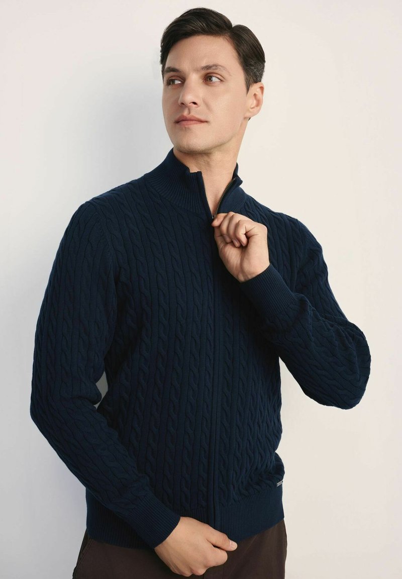 Granatowy sweter zapinany na zamek, pleciony w wzór warkocz, z ryżowymi mankietami i dolnym brzegiem, z wysokim kołnierzem i gładką fakturą.