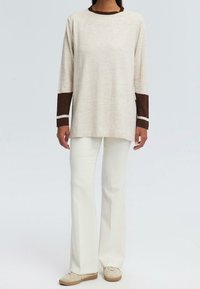 Top beige a maniche lunghe con dettagli marroni ai polsini e al colletto, abbinato a pantaloni bianchi a zampa d'elefante e scarpe beige. Tessuto morbido.