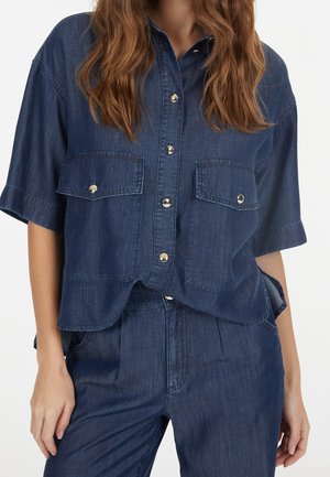 Mujer con camisa de mezclilla azul oscuro de manga corta, abotonada y con bolsillos con solapa en el pecho, combinada con pantalones de mezclilla de talle alto a juego.