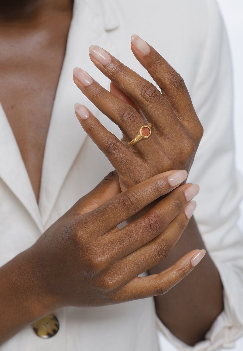 Arany gyűrű kerek vörös kővel, egy jól ápolt, világos rózsaszín körmökkel rendelkező kézen, fehér háttér előtt.