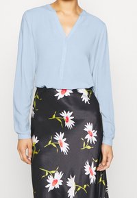 Blouse bleu clair à manches longues avec un col en V et une fermeture à boutons, associée à une jupe noire ornée de grandes marguerites blanches aux centres rouges.
