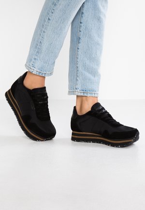 Persona con vaqueros azul claro y zapatillas negras con plataforma y franjas marrones sobre suelo blanco, vista desde la mitad de las pantorrillas hacia abajo.