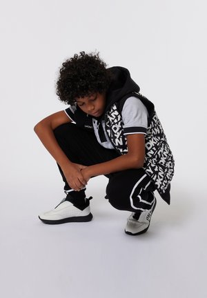 Zwart-witte grafische pufferjack over een grijs poloshirt, gecombineerd met zwarte joggers en witte sportschoenen met zwarte accenten.