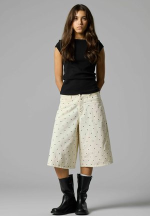 Jeune femme aux longs cheveux bruns portant un haut noir sans manches, un pantalon blanc à jambes larges à pois et des bottes noires mi-mollet, debout devant un fond gris.