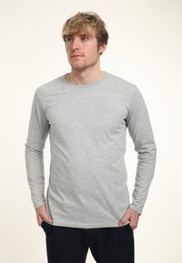 Šedá halenka s dlouhým rukávem a kulatým výstřihem, vyrobená z měkké tkaniny. Obsahuje jednoduchý design, těsný střih a hladkou texturu. Kombinována s tmavými pruhovanými kalhotami.