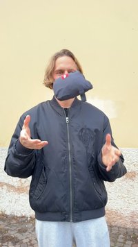 Black bomber jacket with a zip and ribbed cuffs, gray pants, tossing a small, black bag with a red logo, caught in mid-air.