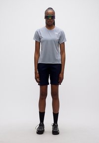 Γυναίκα στέκεται φορώντας γκρι κοντομάνικο μπλουζάκι, μαύρο σορτς, μαύρες κάλτσες, παπούτσια πεζοπορίας και μεγάλα αντανακλαστικά γυαλιά ηλίου μπροστά σε απλό φόντο.