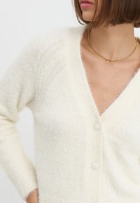 Fluffy oversized crèmekleurige cardigan met een V-hals, knoopsluiting en ribbelmanchetten. Zachte textuur en subtiele stikdetails op de schouders.