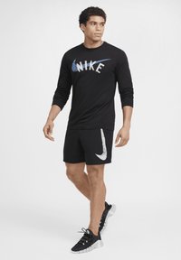 Fekete, hosszú ujjú Nike póló, fehér és kék logóval, fekete rövidnadrággal párosítva, amely fehér akcentussal rendelkezik. Fekete sportcipővel viselve.